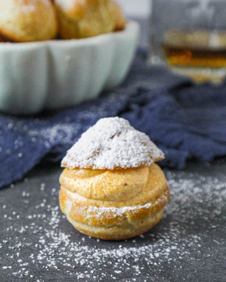 Close-up-of-Boozy-Pumpkin-Cream-Puff-with-bowl-of-unfilled-cream-puffs-and-a-glass-of-whiskey-behind-it.