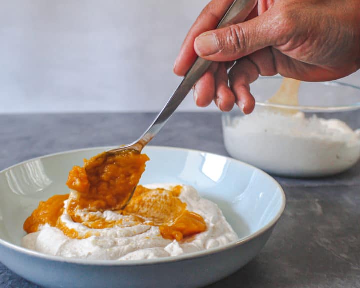  Hand-holding-a-spoon-while-stirring-pumpkin-puree-into-big-blue-bowl-of-plain-whipped-cream-in-front-of-a-clear-bowl-of-plain-whipped-cream.