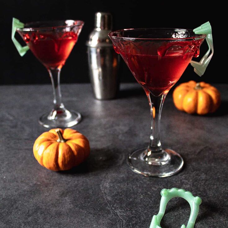 Two Cranberry Orange Martinis on a grey surface with a black background surrounded by small orange pumpkins and glow in the dark vampire teeth. Each martini glass rim has red liquid candy that is dripping down to look like blood.