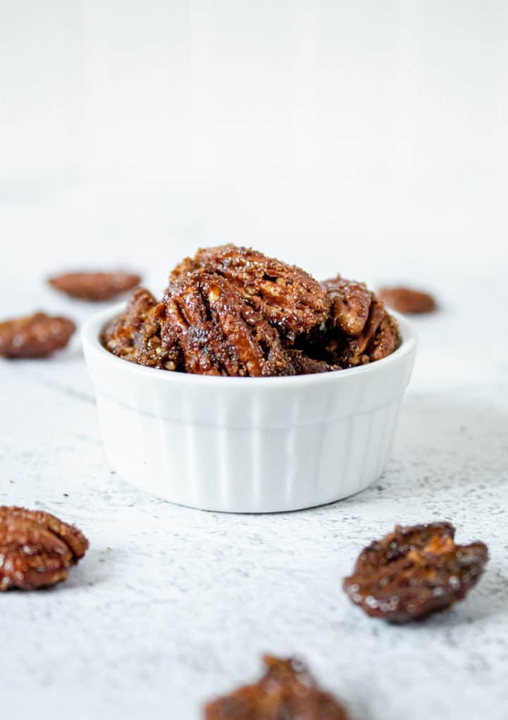 head on shot of candied peacans in small white bowl surrounded by pecans 
