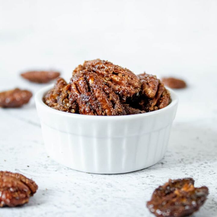 head on shot of candied peacans in small white bowl surrounded by pecans
