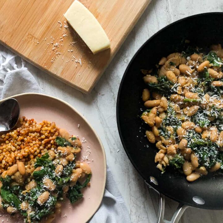 Braised Cannellini Beans And Greens With Lemony Couscous on plate with fork beside cutting board with piece of Parmesan and pan of Braised Cannellini Beans And Greens With Lemony Couscous