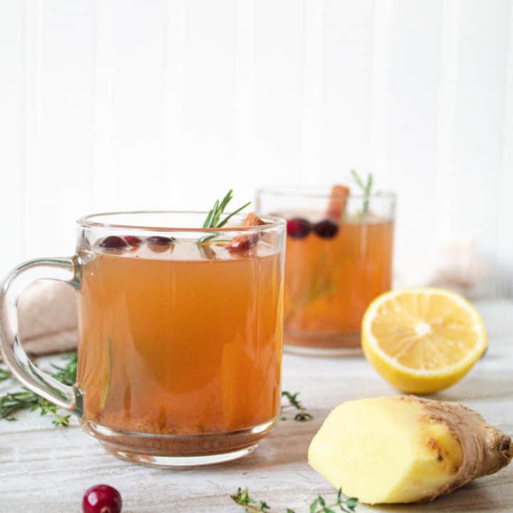 2 glass mugs of herbed mullled cider surrounded by pieces of lemon, ginger and cranberry