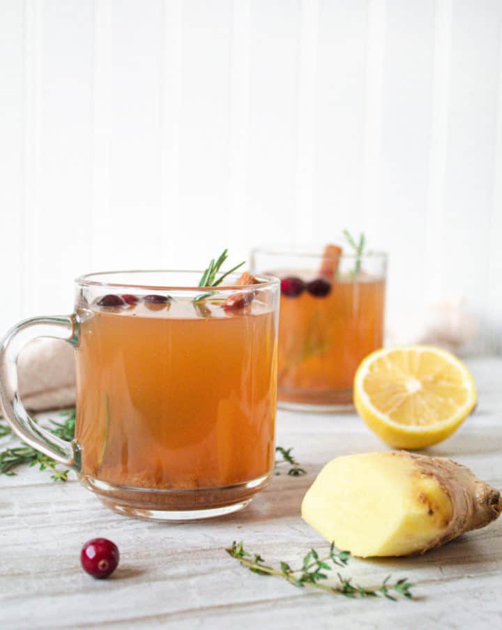 2 glass mugs of herbed mullled cider surrounded by pieces of lemon, ginger and cranberry