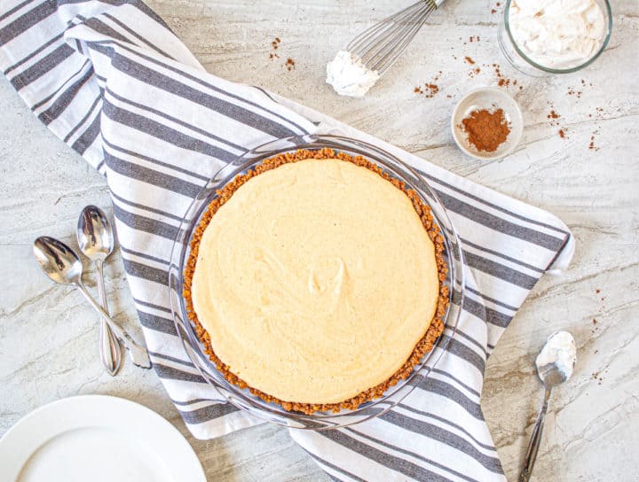 Pumpkin Ice Cream Pie in pie dish on blue stripped cloth surrounded by spoons and ingredients