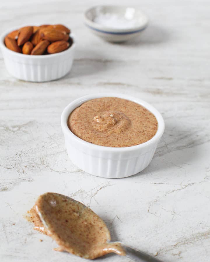 close up of almond butter in bowl next to bowl of almonds and spoon