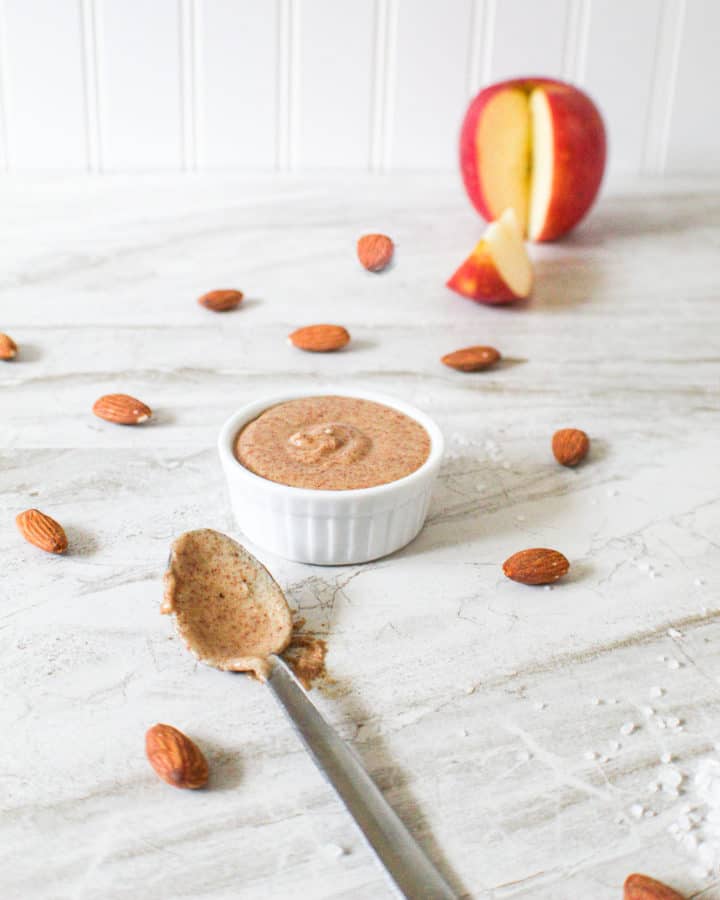 almond butter in small bowl surrounded by almonds on table next to a spoon and a sliced apple