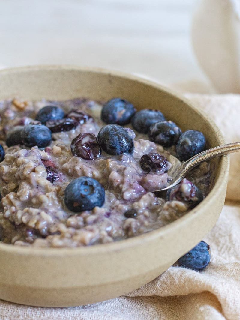 Close up of high protein blueberry oatmeal in a tan bowl topped with fresh blueberries. There is a spoon coming out of the right of the bowl. 