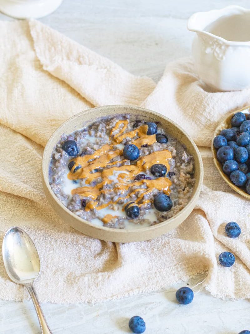 Vegan High Protein Blueberry Oatmeal in a tan bowl topped with extra almond milk, fresh blueberries and almond butter drizzled on top. Oatmeal is sitting on a tan napkin and surrounded by a bowl of blueberries to the right, a silver spoon in the bottom left and a cream holder in the top right corner.