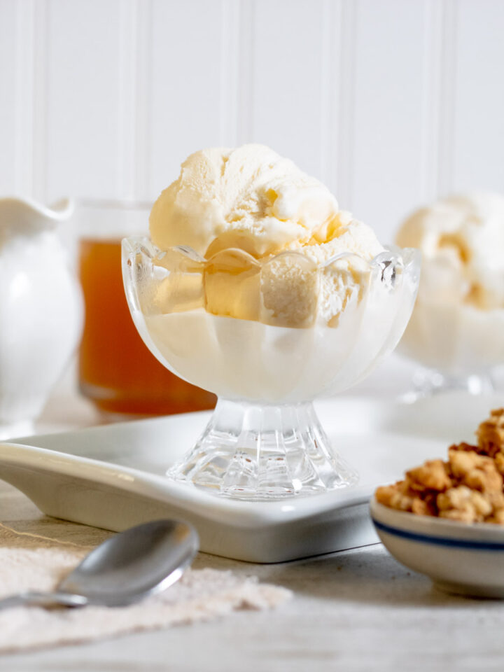 Straight on shot of Vanilla ice cream in a a clear bowl. Bowl is on top of a small white porcelain tray in front of apple cider, another bowl of ice cream, and in the foreground there's on the right side granola and a silver spoon on the left side.
