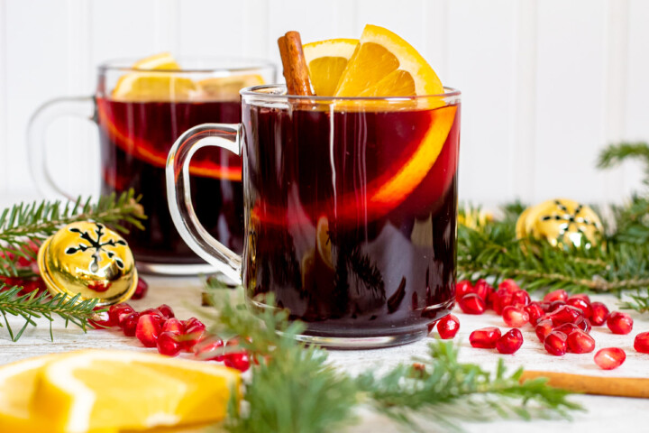 Two clear glass mugs of non-alcoholic mulled wine mocktail with orange slices and a cinnamon stick sticking out on a white surface and background, surrounded by orange slices, Christmas tree branches, pomegranate seeds and jingle bells.