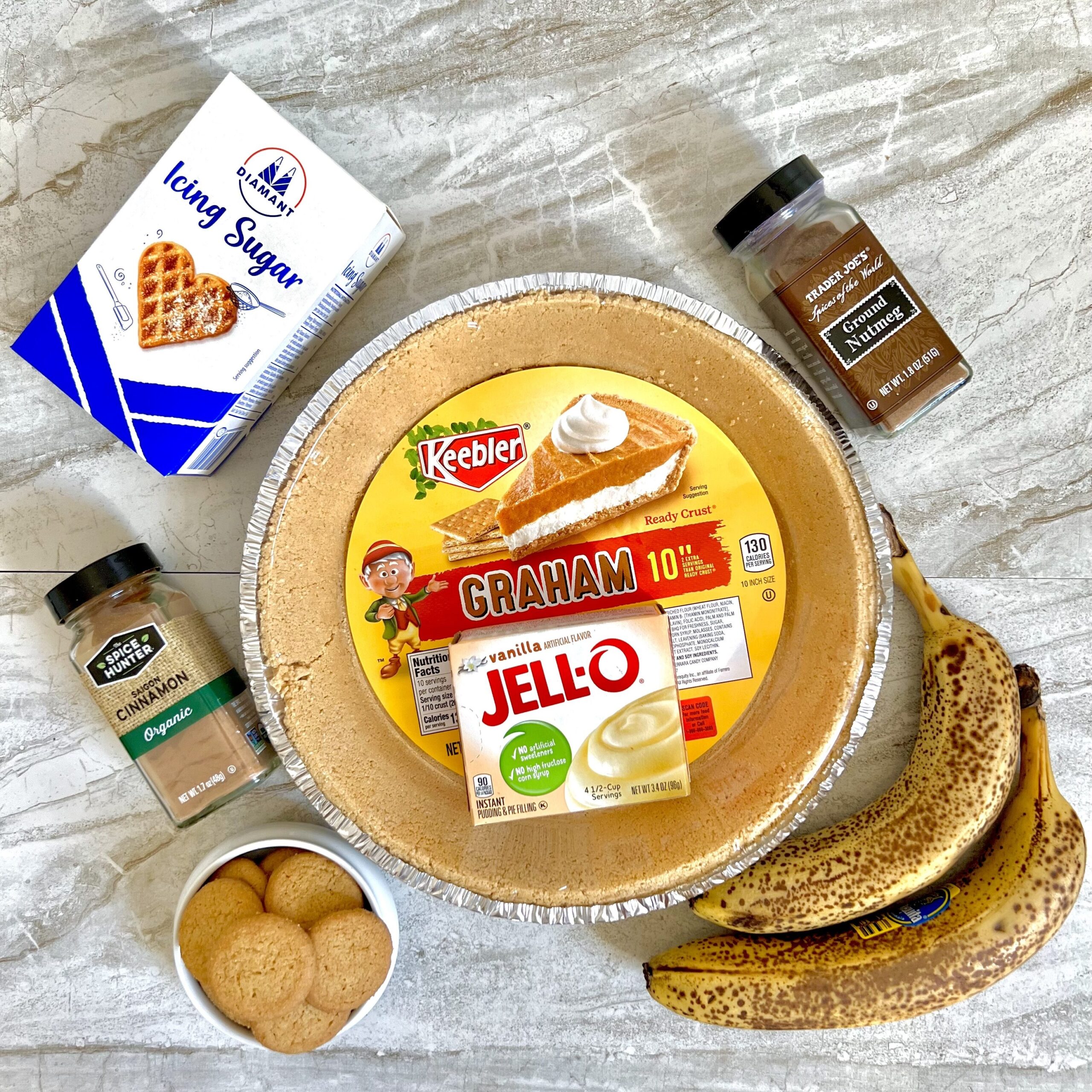 Banana Pudding Pie Ingredients (graham cracker crust in a pie tin with a plastic labeled cover, a box of Jell-o vanilla instant pudding inside the pie crust. The pie crust is surrounded by a bottle of nutmeg, two very ripe bananas, a small bowl of Nila wafers, a bottle of cinnamon and a box of icing sugar.