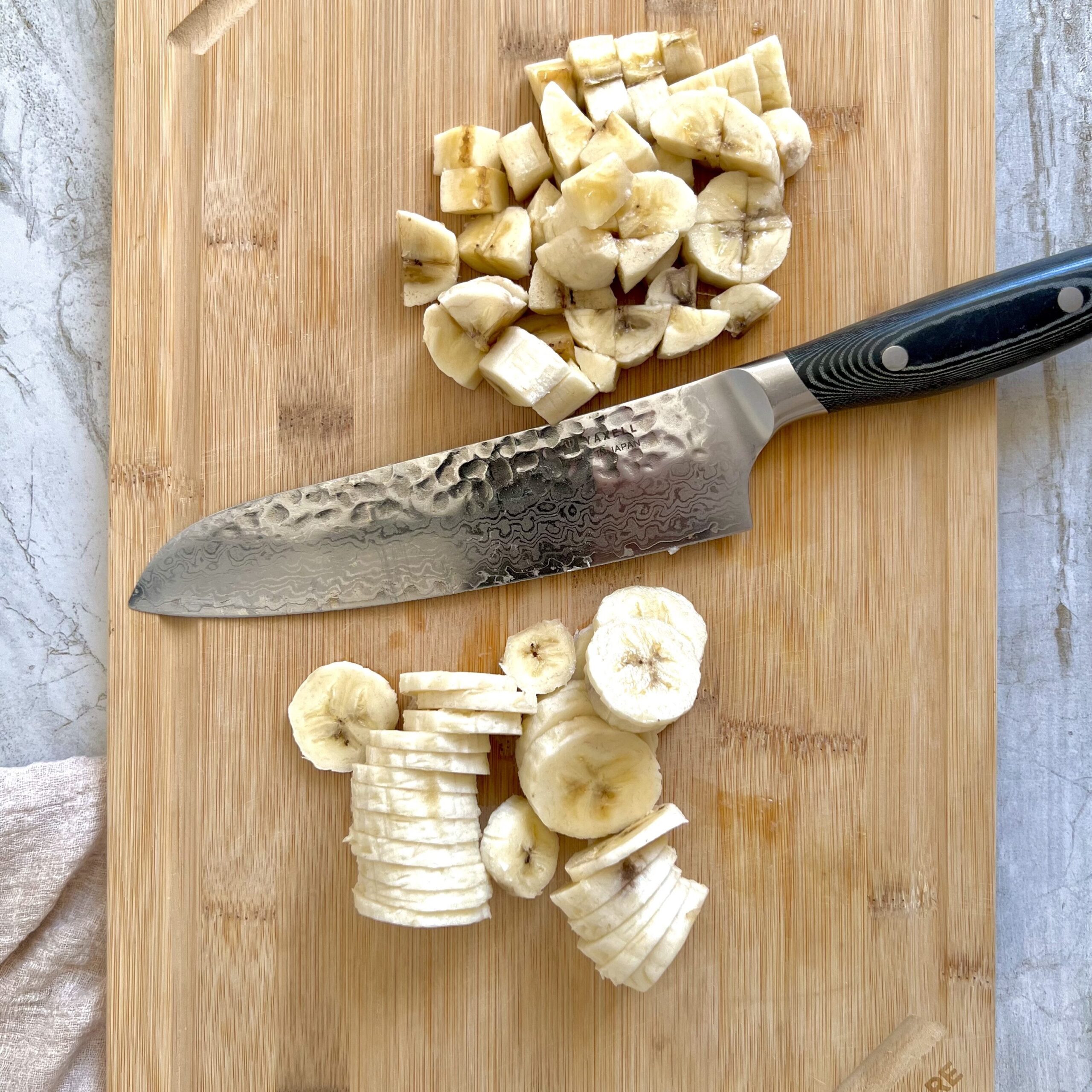 A large knife sitting in the middle of a bamboo cutting board with thinly sliced bananas on one side and pieces of minced bananas on the other side.