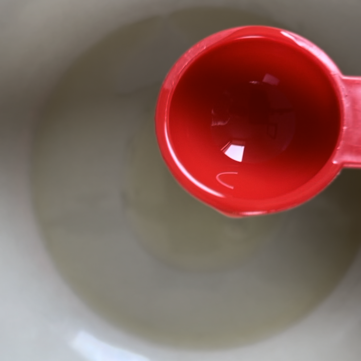 Close up of a red measuring spoon with olive oil in it over a large white ceramic pot. 