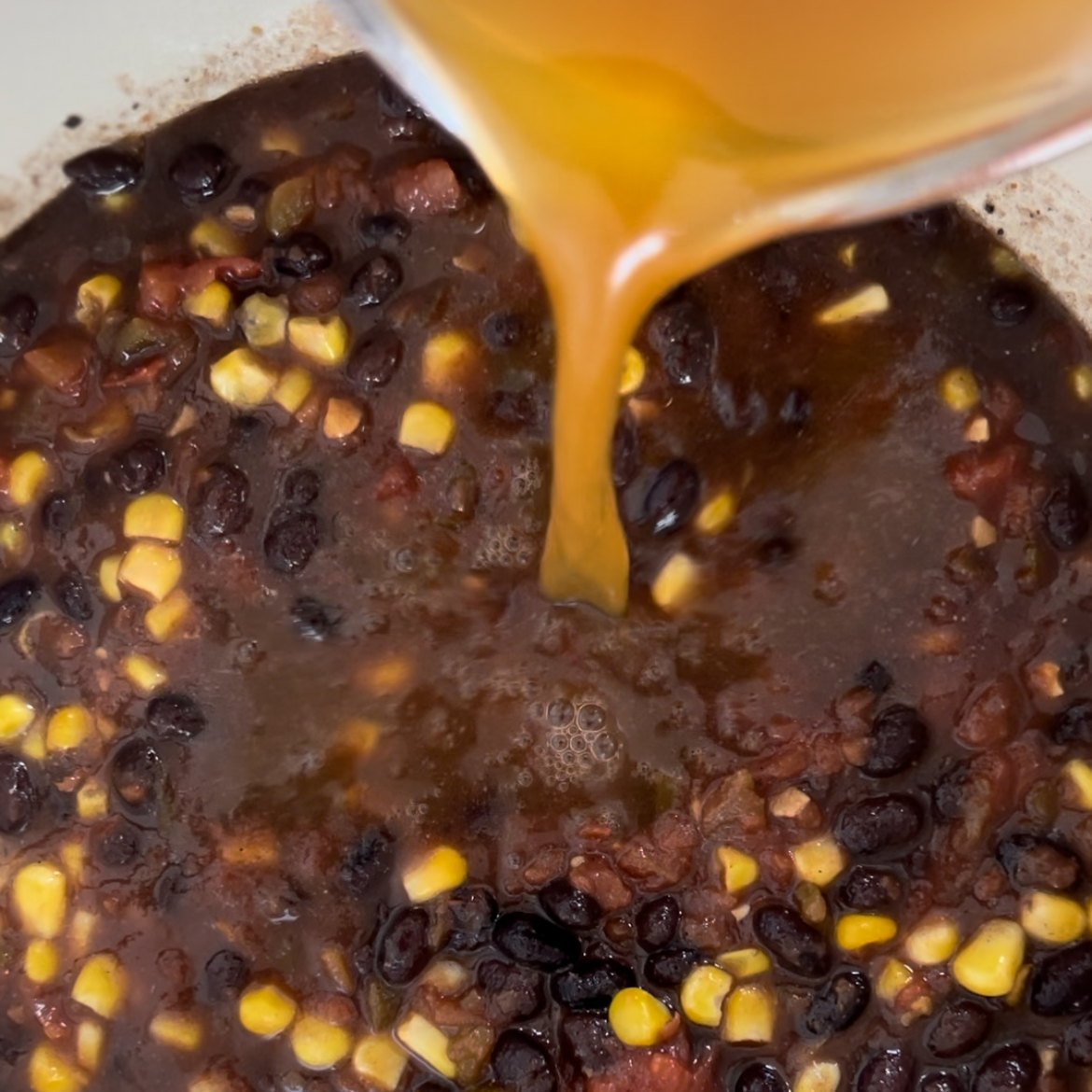 Closeup of veggie broth being poured into a large white pot of easy black bean chili.